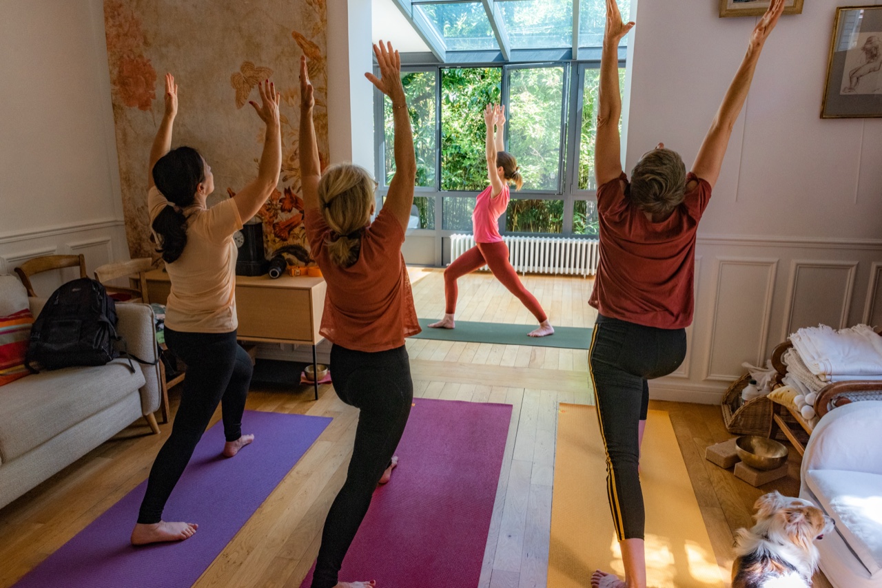 Cours collectif de yoga à Viroflay : quatre élèves en salut au soleil, bras levés, face à la verrière